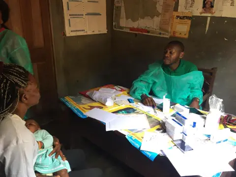 Une maman et son bébé reçus par un soignant derrière un bureau couvert de dossiers.