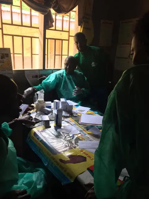 Une soignante en blouse verte prépare des médicaments sur une table de consultation.