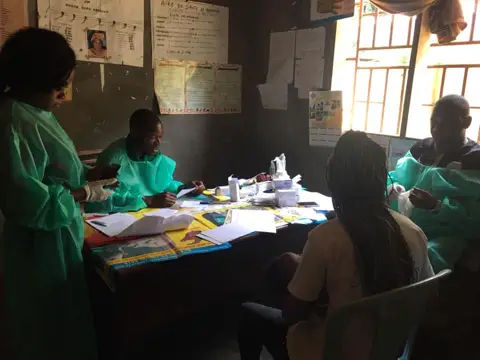Des soignants en blouse verte reçoivent une patiente à une table couverte de médicaments.