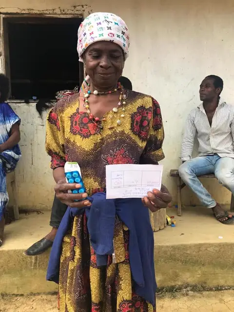 Une habitante en robe à motifs tient ses médicaments et son ordonnance devant le centre de santé.